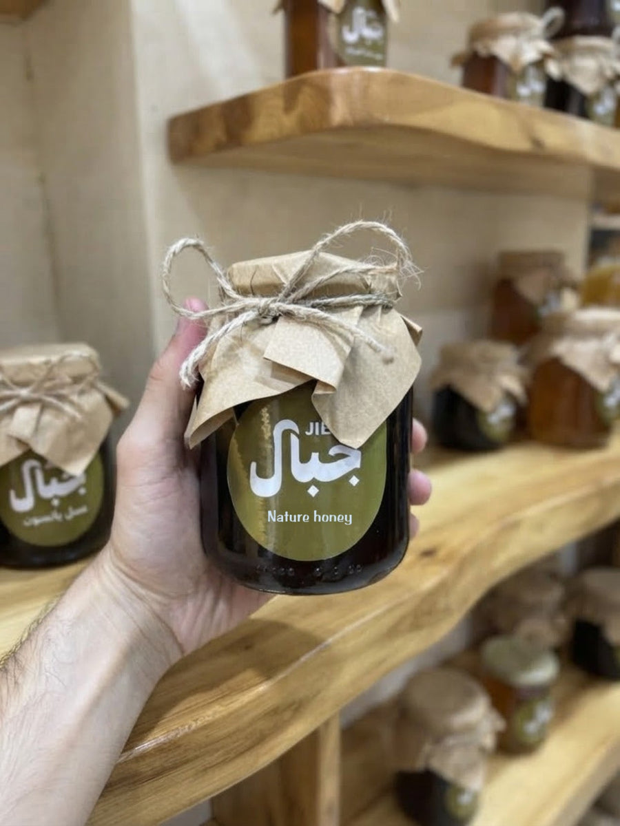 يد تحمل برطمان عسل حبة البركة الداكن من جِبال أمام رفوف خشبية عليها برطمانات عسل