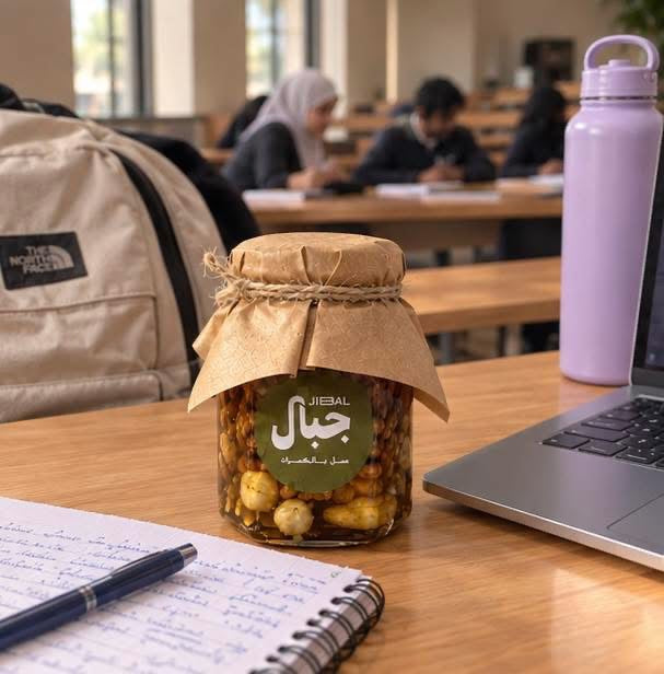 برطمان مكسرات بالعسل من جِبال على مكتب دراسة بجوار حاسوب محمول ودفتر وزجاجة مياه